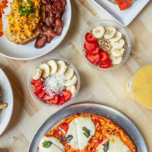 An overhead view of various dishes: pasta, pizza, fruit bowls, soup, chicken wings, fries, and a smoothie on a wooden table.