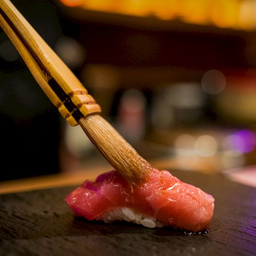 A hand is brushing sauce over a piece of nigiri sushi on a dark surface, with a blurred background suggesting a restaurant setting.