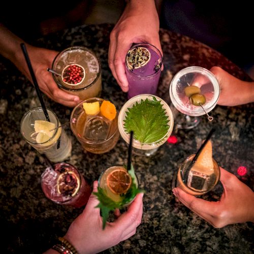 A group of people holding a variety of colorful cocktails with garnishes, shot from above against a dark background.