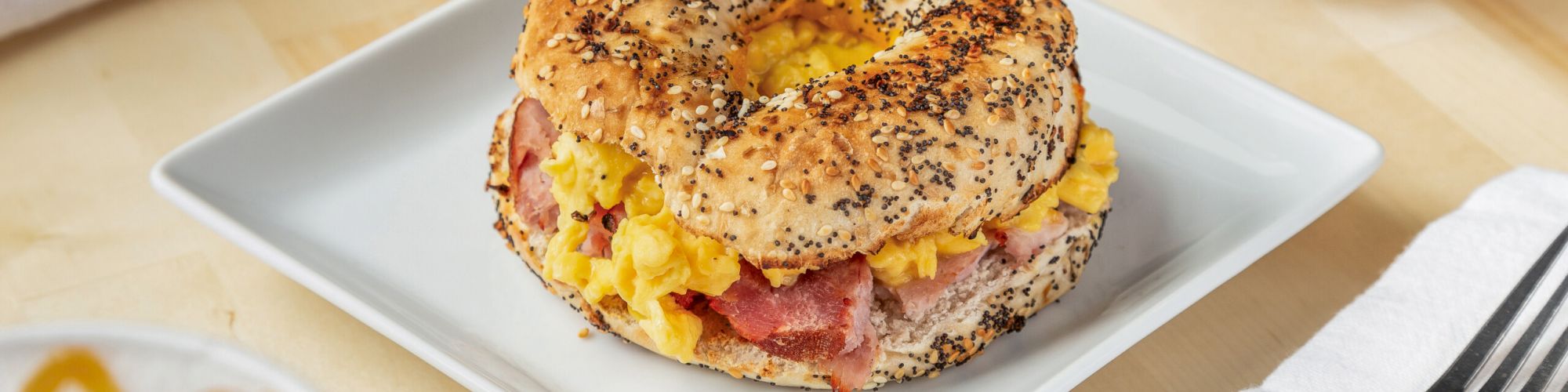 The image shows a bagel sandwich with scrambled eggs and bacon on a white plate, accompanied by a chocolate drink and a yogurt parfait.