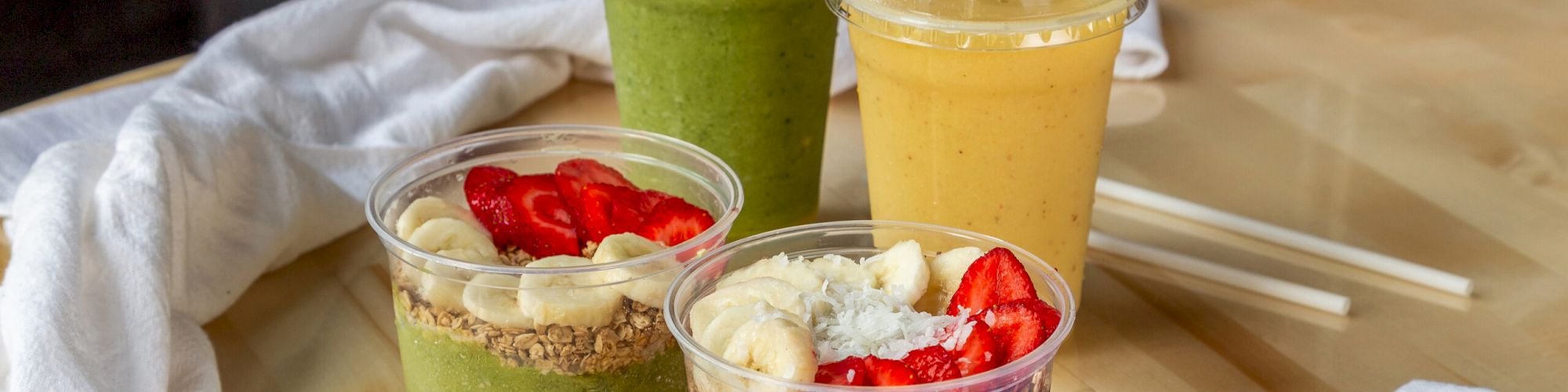 The image shows two smoothie bowls with fruit and granola toppings, and two smoothies, one green and one yellow, on a wooden table with utensils.