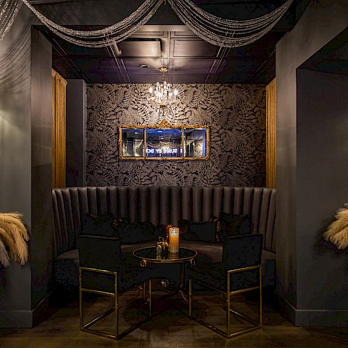 An elegant, dimly lit lounge with dark walls features feathered decor, a tufted banquette, a chandelier, and a small table with chairs underneath a mirror.