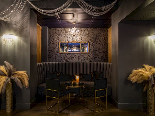 An elegant, dimly lit lounge with dark walls features feathered decor, a tufted banquette, a chandelier, and a small table with chairs underneath a mirror.