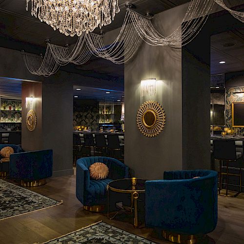 A stylish lounge with elegant chandeliers, blue velvet chairs, ornate mirrors, and a cozy bar area creates a luxurious ambiance.