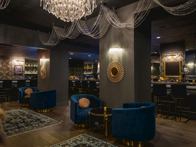 A stylish lounge with elegant chandeliers, blue velvet chairs, ornate mirrors, and a cozy bar area creates a luxurious ambiance.