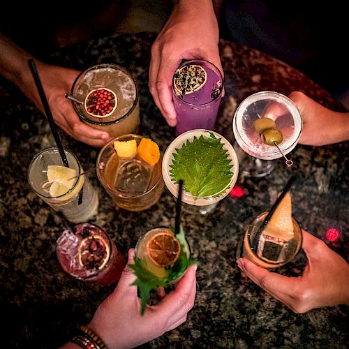 A group of people holding a variety of colorful cocktails with garnishes, viewed from above, on a dark, speckled surface.