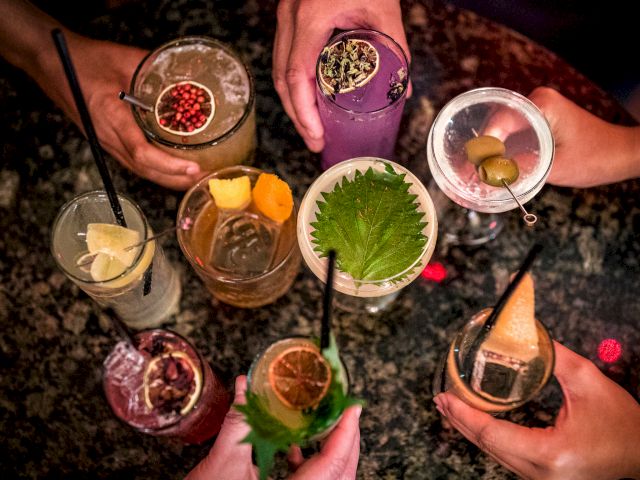 A group of people holding a variety of colorful cocktails with garnishes, viewed from above, on a dark, speckled surface.