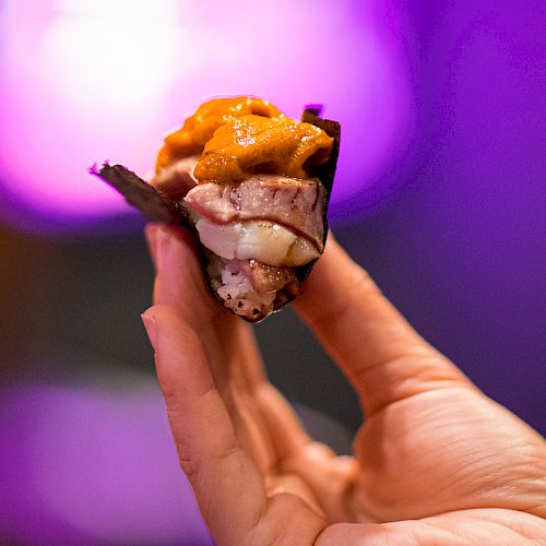 A hand holds a piece of sushi with vibrant, colorful toppings against a blurred, purple-lit background.