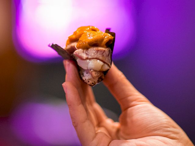 A hand holds a piece of sushi with vibrant, colorful toppings against a blurred, purple-lit background.