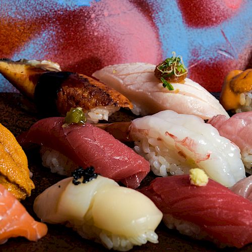 An assortment of nigiri sushi with various types of fish and seafood, presented on a wooden tray, against a colorful, artistic backdrop.