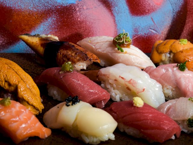 An assortment of nigiri sushi with various types of fish and seafood, presented on a wooden tray, against a colorful, artistic backdrop.