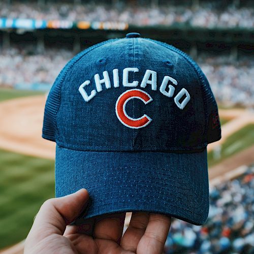 The image shows a hand holding a blue baseball cap with "Chicago C" on it at a stadium. The field and a crowd are visible in the background.