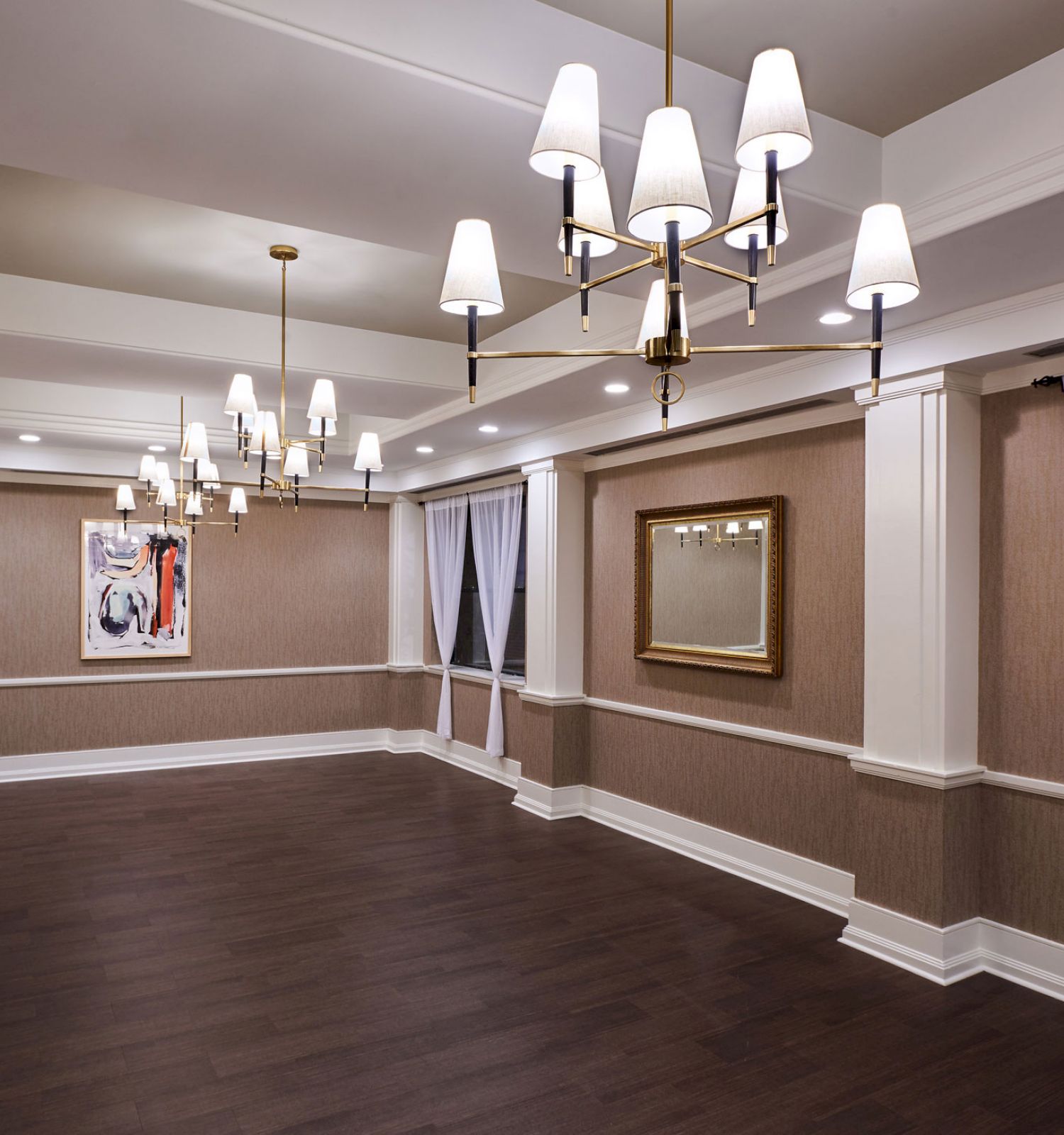 A spacious, elegant room with polished dark wood floors, chandeliers, framed artwork, curtains on windows, and recessed lights in the ceiling.