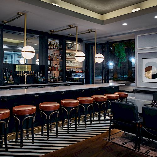 This image shows a stylish bar with a long counter, leather stools, modern lighting, a stocked shelf of bottles, and contemporary wall art.