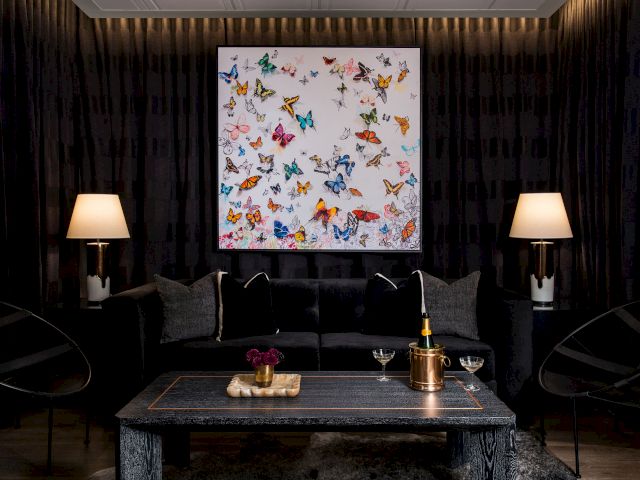 A stylish, dark-toned living room with a black sofa, two lamps, a coffee table, and a colorful butterfly artwork on the wall, creating a sophisticated ambiance.