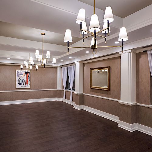 A spacious, empty room with dark hardwood floors, elegant chandeliers, framed artwork, large curtains on windows, and recessed ceiling lights.