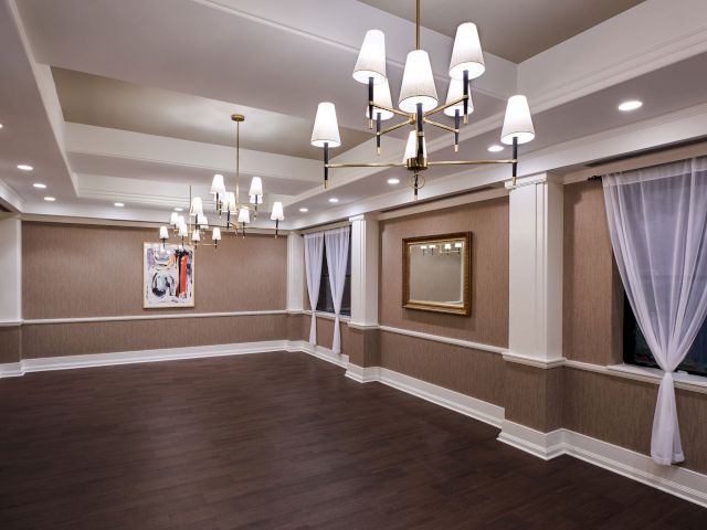 A spacious, empty room with dark hardwood floors, elegant chandeliers, framed artwork, large curtains on windows, and recessed ceiling lights.