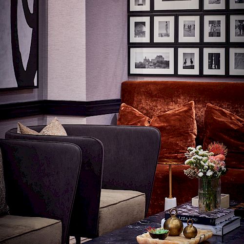 A cozy living room with a black marble coffee table, flowers, pears, books, a red velvet couch, dark armchairs, framed photos, and wall art.