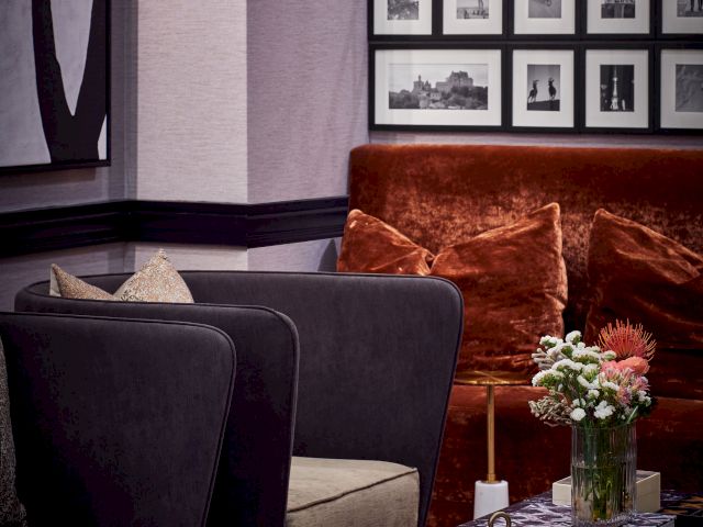 A cozy living room with a black marble coffee table, flowers, pears, books, a red velvet couch, dark armchairs, framed photos, and wall art.