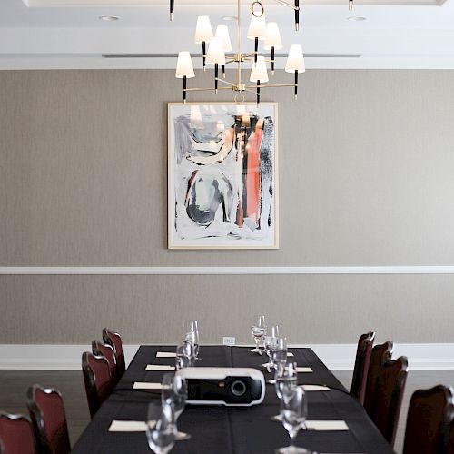 A long table is set with glasses and napkins in a modern meeting room. A projector is on the table, and abstract art hangs on the wall.