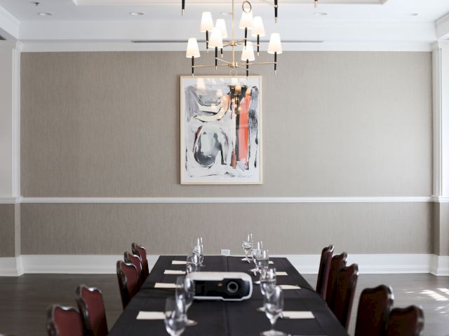 A long table is set with glasses and napkins in a modern meeting room. A projector is on the table, and abstract art hangs on the wall.