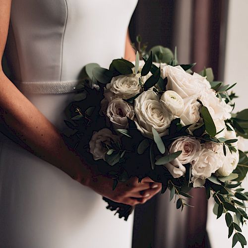 A person is holding a bouquet of white and light pink roses with greenery, wearing a white outfit, likely at a wedding.