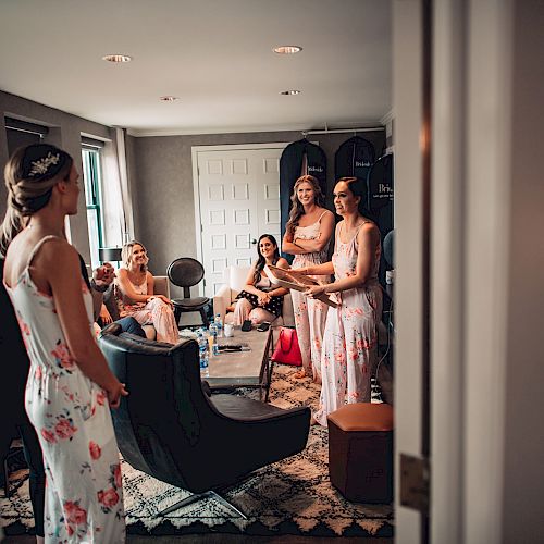 A group of women in floral dresses are gathered in a room, chatting and laughing, possibly preparing for an event or celebration.