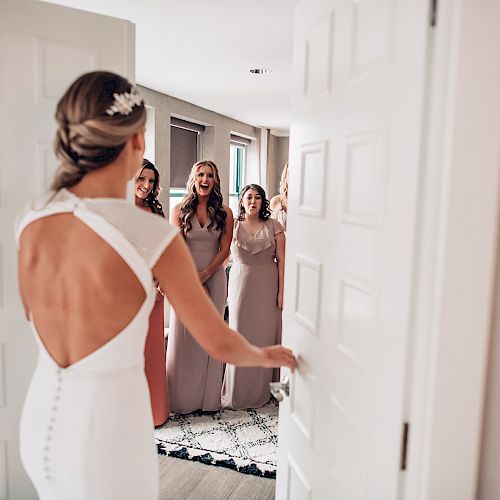 A bride in a white dress opens a door, revealing three women in bridesmaid dresses who appear excited and happy to see her.