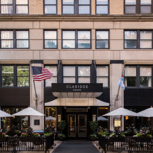 The image shows the entrance of Claridge House with two flags, a seating area with white umbrellas, and greenery surrounding the entrance.