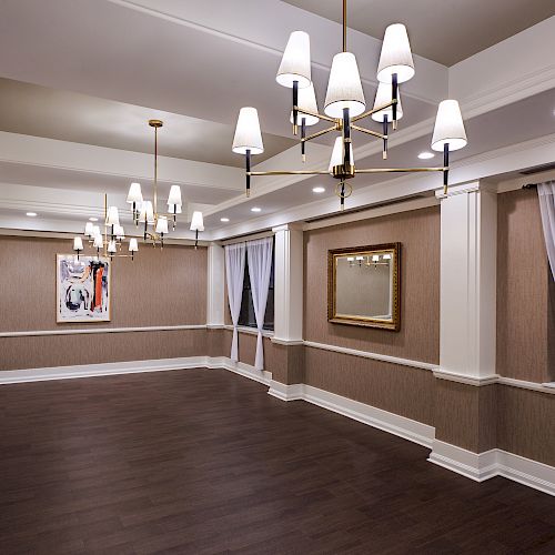 This image shows an elegant, empty room with dark wooden flooring, two chandelier fixtures, framed paintings, and large windows with white curtains.