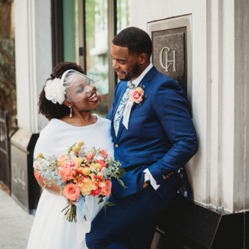 A bride holding a bouquet and a groom in a blue suit share a joyful moment outside, standing against a building facade.