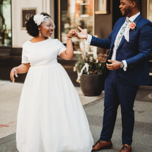 A happy couple dressed in wedding attire is dancing on a city street, with the bride in a white gown and the groom in a blue suit.