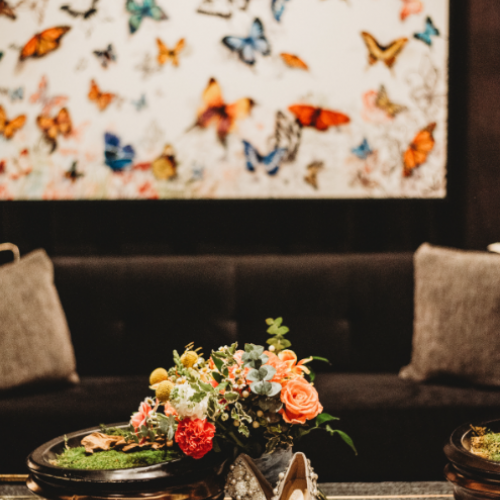 A vibrant arrangement of flowers and a pair of elegant shoes set on a table, with a butterfly-themed artwork in the background.