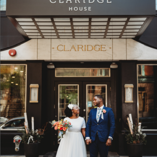 A newlywed couple is posing in front of the Claridge House entrance. The bride is in a white dress and the groom is in a blue suit.