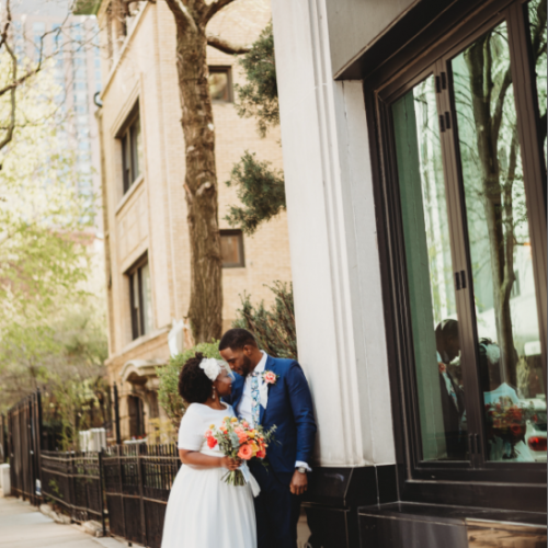 A bride and groom stand closely on a city sidewalk, with the bride holding a colorful bouquet. They are near a building with a "C1" sign outside.