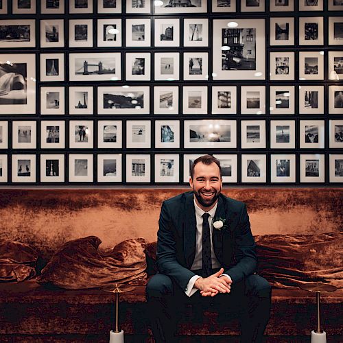 A man in a suit is sitting on a brown couch in front of a wall filled with many framed photographs, with wall sconces on either side of the display.