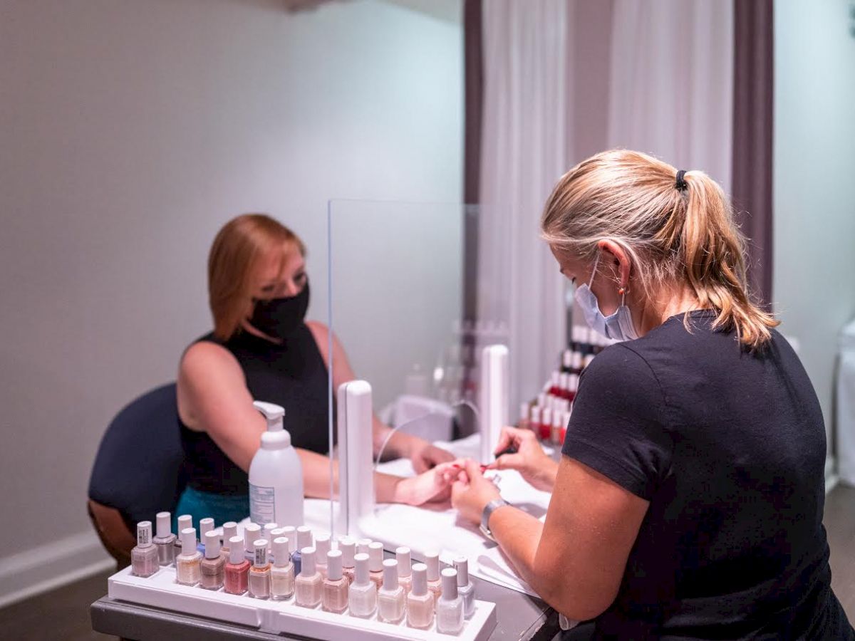 A woman is getting her nails done at a salon, both individuals are wearing masks. Various nail polish bottles are displayed in the foreground.