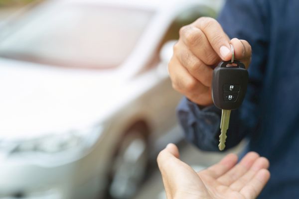 A hand is holding a car key fob above another person's open hand, with a blurred car in the background.