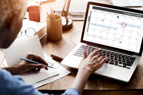 A person is working at a desk with a laptop displaying a calendar planner, while taking notes in a notebook.