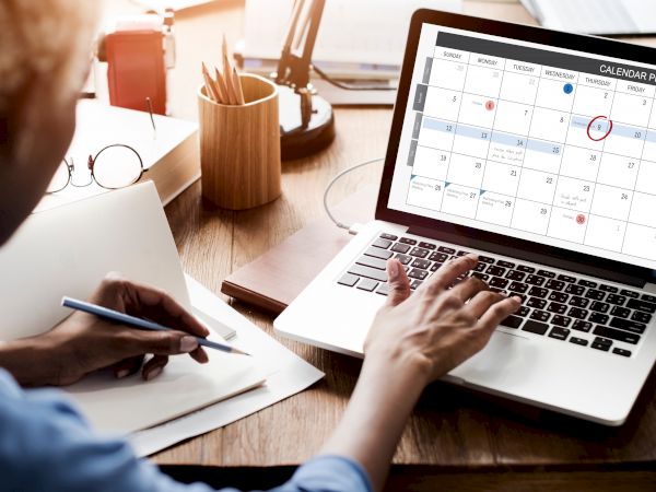A person is working at a desk with a laptop displaying a calendar planner, while taking notes in a notebook.