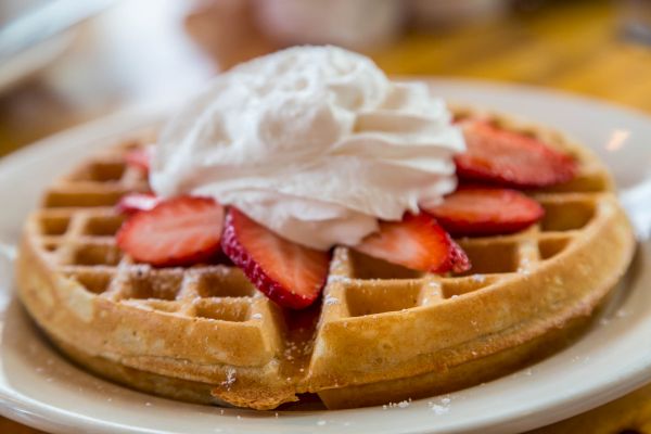 A golden waffle topped with sliced strawberries and a dollop of whipped cream is on a white plate, ready to be enjoyed.
