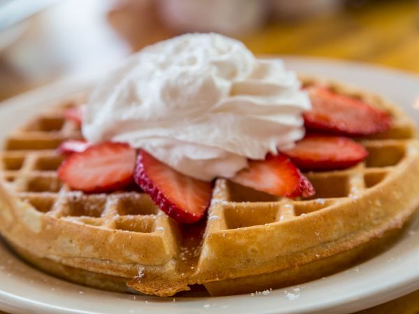 A golden waffle topped with sliced strawberries and a dollop of whipped cream is on a white plate, ready to be enjoyed.