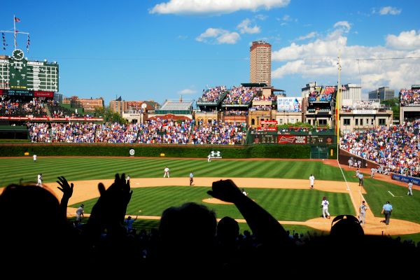 The image shows a baseball stadium during a game, with fans cheering and players on the field. There are clear skies and a tall building in the background.