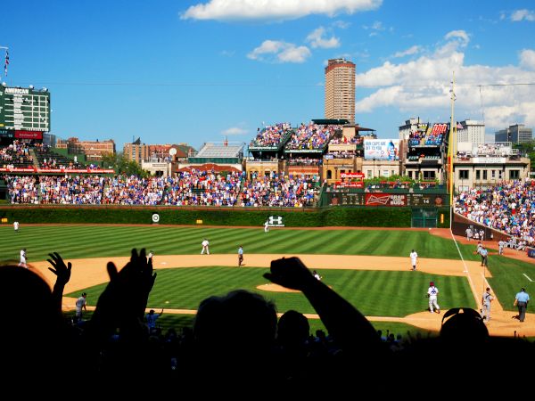 The image shows a baseball stadium during a game, with fans cheering and players on the field. There are clear skies and a tall building in the background.