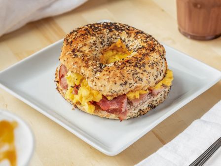 A bagel sandwich with scrambled eggs and bacon on a square white plate, with a fork and napkin nearby, and a drink and dessert in the background.