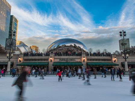 People ice skating in an outdoor rink with a reflective sculpture and city buildings in the background under a blue sky.