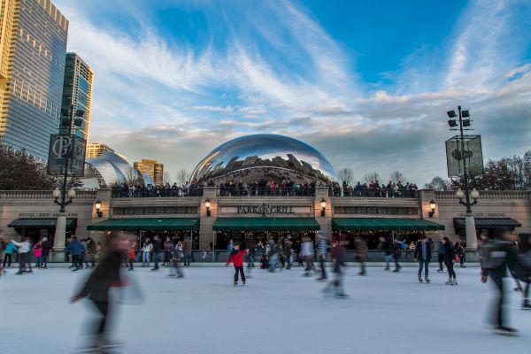 People ice skating in an outdoor rink with a reflective sculpture and city buildings in the background under a blue sky.