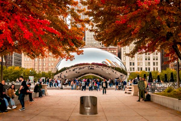 People are gathered around the Cloud Gate (The Bean) sculpture in Chicago's Millennium Park, framed by vibrant autumn foliage and city buildings.