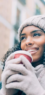 A person in warm clothing, including a knit hat and gloves, is holding a red cup and smiling on a city street with blurred buildings in the background.