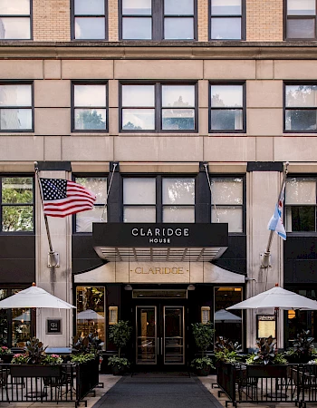 The image shows the exterior of Claridge House, featuring American and Chicago flags, with outdoor seating and umbrellas in front.